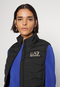 Črn prešite brezrokavnik z visokim ovratnikom, z zlatim logotipom "EA7 Emporio Armani". Nosi se preko modre majice. Gladka tekstura. Zadrga spredaj.