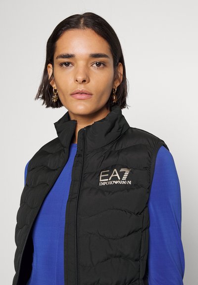 Черен ватиран елек с висока яка, украсен с логото "EA7 Emporio Armani" в златисто. Носен върху син топ. Гладка текстура. С цип отпред.