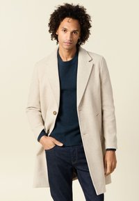 Abrigo de lana beige con solapas de pico, cierre de un solo botón y dos bolsillos frontales, llevado sobre un suéter oscuro y jeans. Textura suave.