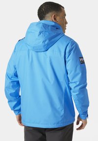 Blauwe waterdichte jas met een capuchon, voorzien van elastische manchetten en een logo op de linker mouw. Soepele textuur en gestroomlijnd ontwerp.