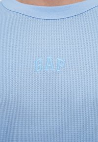 Ανοιχτό μπλε, υφασμάτινο κοντομάνικο πουκάμισο με στρογγυλή λαιμόκοψη και κεντημένο λογότυπο "GAP" στο ίδιο μπλε χρώμα στην περιοχή του στήθους.