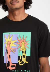 Svart bomulls t-skjorte med et fargerikt grafisk motiv av to stiliserte katter mot en pastellbakgrunn med stjerneformede figurer og "Volcom" tekst.