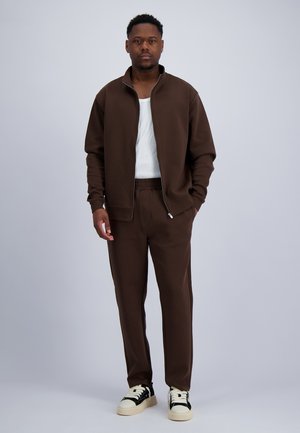 Mann steht, trägt eine braune Zip-up-Jacke, passende Hose, weißes Hemd und schwarz-weiße Sneakers vor einfarbigem Hintergrund.