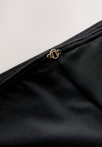 Közeli kép fekete anyagról, varrott széllel és kis kör alakú arany kapoccsal, amely minimalista geometriai mintát ábrázol.