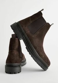 Botine din suede maro, cu laturi elastice, suprafață texturată și talpă din cauciuc negru cu dâre adânci pentru aderență. Buclă pentru a fi ușor de încălțat.