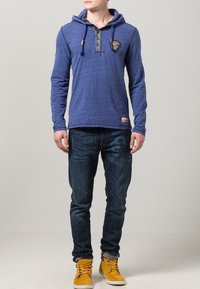 Felpa blu a maniche lunghe con tasca frontale, colletto con bottoni e toppa del logo, abbinata a jeans di denim scuro e scarpe da ginnastica gialle.