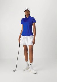 Μπλε πόλο μπλούζα με κοντά μανίκια, λευκή πλισέ φούστα και λευκά παπούτσια γκολφ. Στεκόμενη με ένα γκλοφ και φοράει λευκό καπέλο.