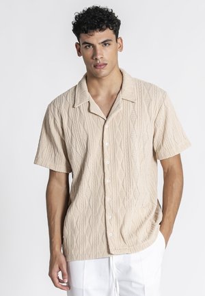 Mężczyzna o krótkich kręconych włosach, ubrany w beżową, teksturowaną, krótkim rękawem koszulę na guziki oraz białe spodnie, stojący z jedną ręką w kieszeni.