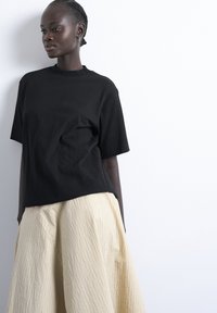 Femme aux cheveux tressés portant un t-shirt noir ample et une large jupe beige, debout contre un mur blanc, regardant sur le côté.