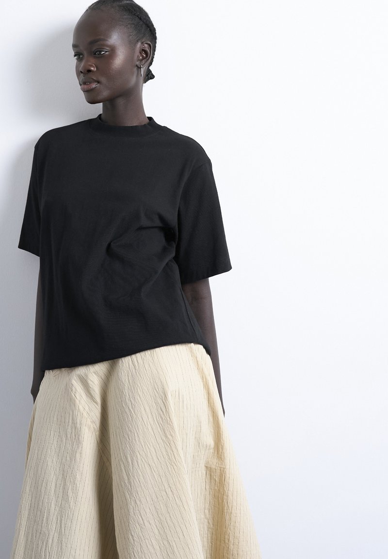 Femme aux cheveux tressés portant un t-shirt noir ample et une large jupe beige, debout contre un mur blanc, regardant sur le côté.