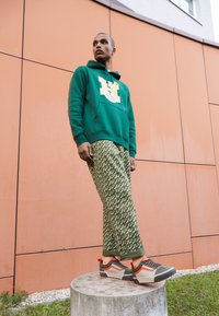 Grön hoodie med en stor grafisk logotyp, mönstrade gröna byxor och grå-orange sneakers, stående på en betongpelare mot en brun vägg.