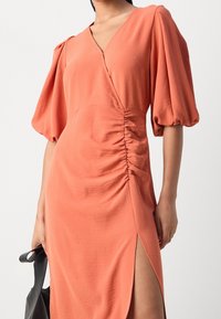 Robe en terre cuite avec une silhouette ajustée, des manches bouffantes, un décolleté en V profond, des fronces sur les côtés et une fente jusqu'à la cuisse. Texture lisse et tissu doux.