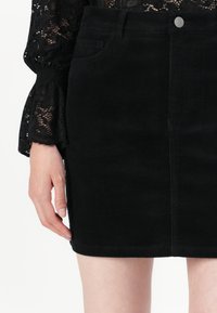 Mini-jupe en velours côtelé noire portée avec un haut en dentelle noire à manches longues, montrant une poche avant et une fermeture par bouton en métal.