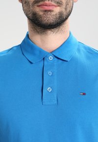 Polo en coton bleu avec une finition texturée, une patte de boutonnage à trois boutons et un petit logo aux couleurs rouge, blanc et bleu sur la poitrine.