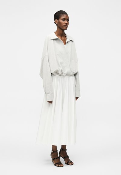 Γυναίκα που φοράει ένα ανοιχτό γκρι φαρδύ φούτερ με γιακά, λευκή μίντι φούστα και μαύρα πέδιλα με λουράκια και τακούνια, στέκεται μπροστά σε απλό φόντο.