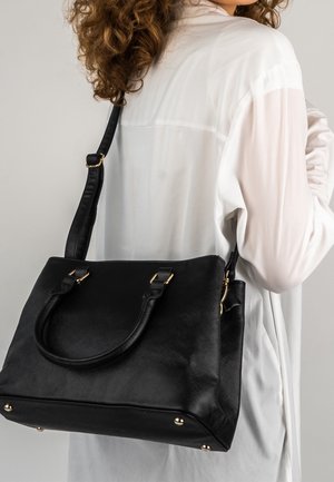 Femme en blouse blanche portant un sac à main en cuir noir texturé avec des ferrures dorées sur l'épaule, sur fond blanc.