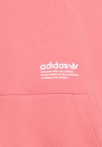 Sudadera rosa de Adidas con un bolsillo frontal. Presenta texto del logo en blanco y el logo de las tres bandas. Hecha de material suave con una textura lisa.