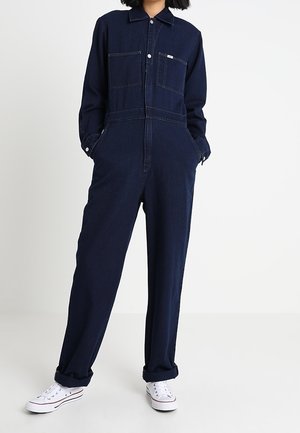 Granatowy kombinezon jeansowy z długimi rękawami, zapinany z przodu na guziki, z dwoma kieszeniami na piersi i luźnym krojem. Noszony z białymi trampkami.