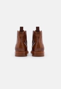 Hnědé kožené kotníkové boty s brogue detaily, elastickými bočními panely a béžovým dřevěným podpatkem. Hladký povrch s perforovanými akcenty.