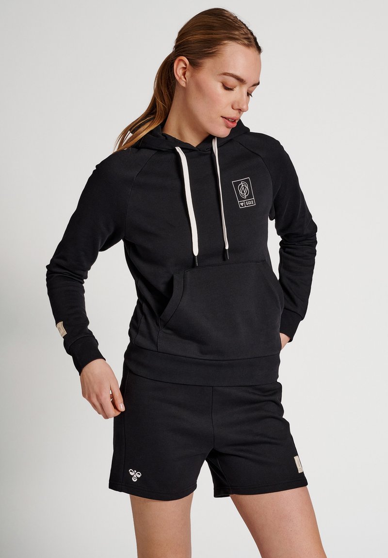 Kobieta ubrana w czarną bluzę z kapturem z białymi troczkami i dopasowane czarne szorty, stoi z jedną ręką w kieszeni na jednolitym tle.
