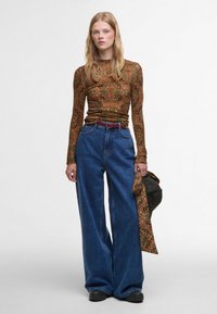 Lange mouwen, bruine patroon top gecombineerd met wijde blauwe denim jeans. Accentriem in bordeaux, zwarte schoenen, met een groene tas.