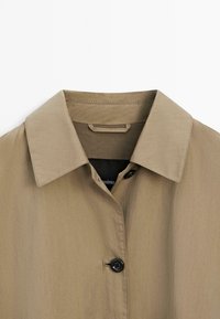 Beige Hemd mit Knopfverschluss, das einen spitzen Kragen, ein leichtes Material und einen einzelnen schwarzen Knopfdetails auf der Vorderseite aufweist.