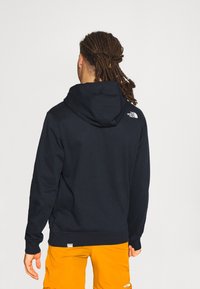 Mu muž s vázanými dredy oblečený v černé mikině a hořčičně žlutých šortkách stojící zády k jednoduchému bílému pozadí.