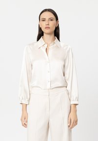 Cream satin blouse with a button-down front, pointed collar, and long sleeves with elastic cuffs, paired with light-colored trousers.