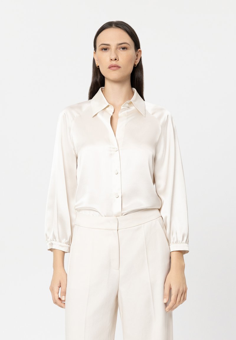 Cream satin blouse with a button-down front, pointed collar, and long sleeves with elastic cuffs, paired with light-colored trousers.