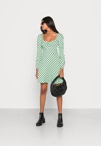 Vestido verde menta com bolinhas pretas, mangas longas, decote quadrado, combinado com uma mala acolchoada preta e botas de tornozelo pretas.