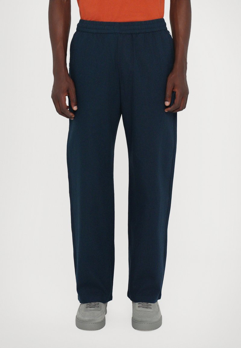 Marineblaue Sweatpants aus weichem Stoff, mit einem elastischen Bund und weiten, geraden Beinen. Kombiniert mit grauen Turnschuhen.