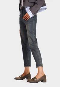 Jeans in denim scuro con taglio affusolato, orlo sfrangiato, abbinati a mocassini con tacco a blocco marroni e dettagli in metallo dorato.