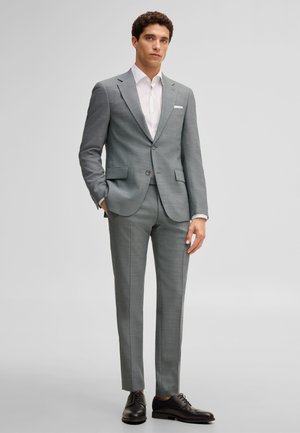 Mężczyzna stoi, ubrany w dopasowany szary garnitur z pasującymi spodniami, białą koszulę, czarne eleganckie buty, ręka w kieszeni, neutralne tło.