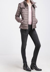 Padded, taupe jacket with a zip front and snap collar, paired with a light pink top, dark jeans, and black ankle boots.