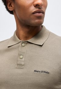Κοντινό πλάνο ενός ατόμου που φοράει μπεζ πικέ πόλο με δύο κουμπιά και κεντημένο το λογότυπο «Marc O’Polo» στο στήθος.