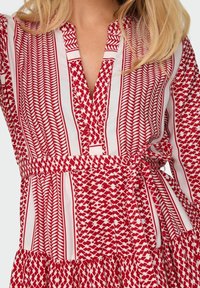 Robe à motifs rouge et blanc avec un décolleté en V profond, un détail de boutons et une taille ceinturée. Comprend des motifs géométriques variés et une jupe évasée.