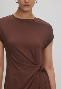 Braunes T-Shirt mit kurzen Ärmeln aus weichem Stoff, das an der Taille mit einem verdrehten Knoten versehen ist und einen einfachen Rundhalsausschnitt hat.