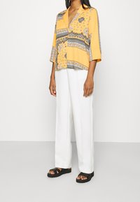 Blusa gialla con motivo e dettagli neri, maniche corte, abbinata a pantaloni bianchi a gamba larga e sandali neri.