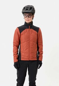 Gewatteerd jack met een roestoranje voorkant, zwarte mouwen, een hoge kraag en een doorgestikt patroon. Draagt handschoenen en een zwart helm.