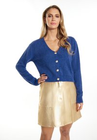 Cardigan blu peloso con scollatura a V e sei bottoni in argento, abbinato a una minigonna dorata metallica con tasche frontali e dettagli di cucitura.