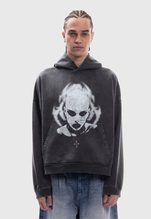 Joven con el pelo trenzado lleva una sudadera negra de gran tamaño con un gráfico de cara de extraterrestre blanca y vaqueros azules sueltos sobre un fondo liso.
