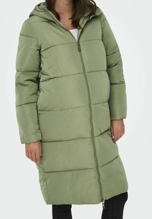 Frau trägt einen langen, wattierten olivgrünen Wintermantel mit Kapuze und Frontreißverschluss, kombiniert mit schwarzen Hosen.