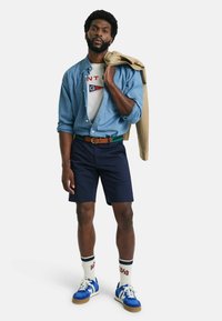 Denimskjorte, hvit grafisk t-skjorte med røde og blå detaljer, marineblå shorts, brun belte, blå joggesko og hvite sokker med rødt trykk.