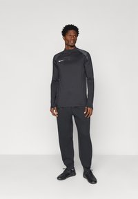 Conjunto atlético preto composto por uma parte superior de manga longa com zipper e detalhes cinzas, acompanhada de calças de treino a condizer; ambos feitos de um tecido leve e texturizado.