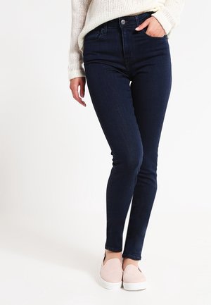 Woman wearing dark blue skinny jeans, white knit sweater, and light pink slip-on shoes, standing with one hand in pocket.