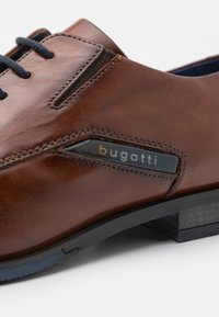 Bruine leren schoen met marineblauwe veters, een slank ontwerp en een zwart accentpaneel met de merknaam "bugatti." Textuurdetails zichtbaar.