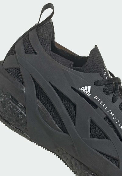 Chaussure de sport noire avec une semelle en caoutchouc texturé, une tige en mesh et des sangles en caoutchouc superposées distinctives. Comprend le logo et le nom de la marque Adidas.