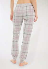 Donna in piedi a piedi nudi che indossa pantaloni del pigiama grigi e rosa a scacchi con cintura elastica, vista da dietro su uno sfondo bianco.