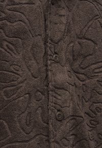 Gros plan d'un tissu texturé marron foncé avec des motifs floraux abstraits en relief et trois boutons noirs cousus en ligne verticale.