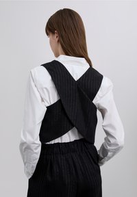 Czarna kamizelka w wzory z tyłem w kształcie litery X, noszona na białej koszuli, wykonana z teksturowanej tkaniny i z dopasowanym paskiem w talii.